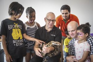 A group of children and adults watching a man holding a snake.