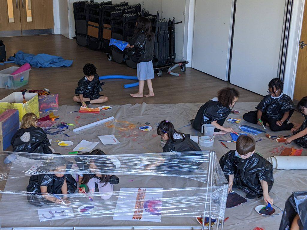 A group of children painting and drawing, on a large mat on the floor.
