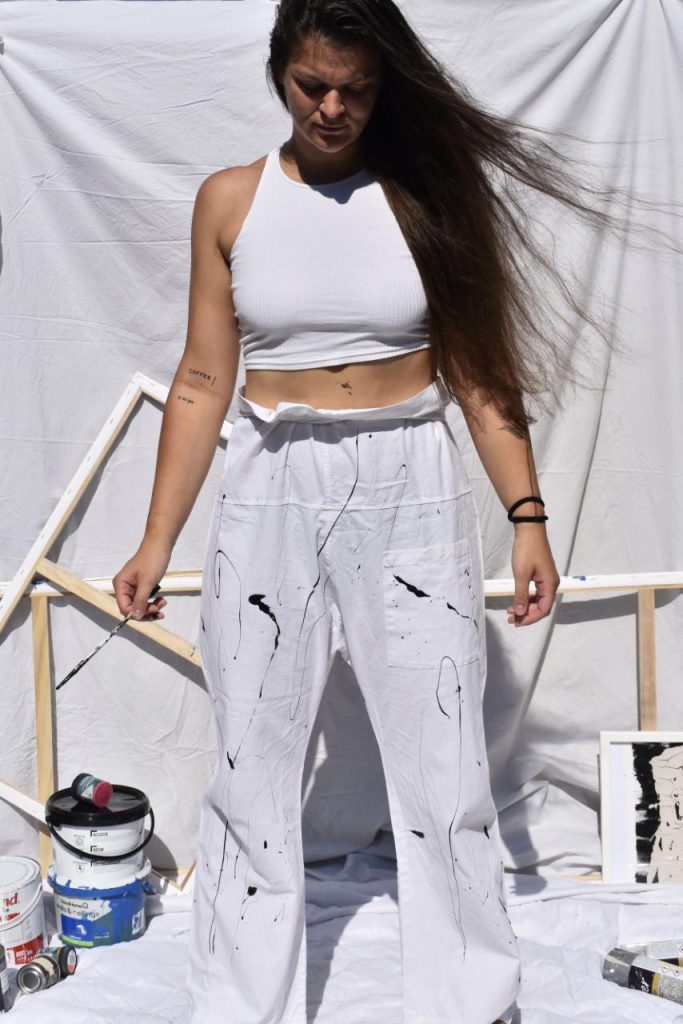 Roberta Brasola, a White, female-presenting person, with long dark hair, is wearing a white top and white trousers, standing in front of a white sheet and canvas frames, surrounded by paint pots. Her clothes have streaks of dark paint on them.