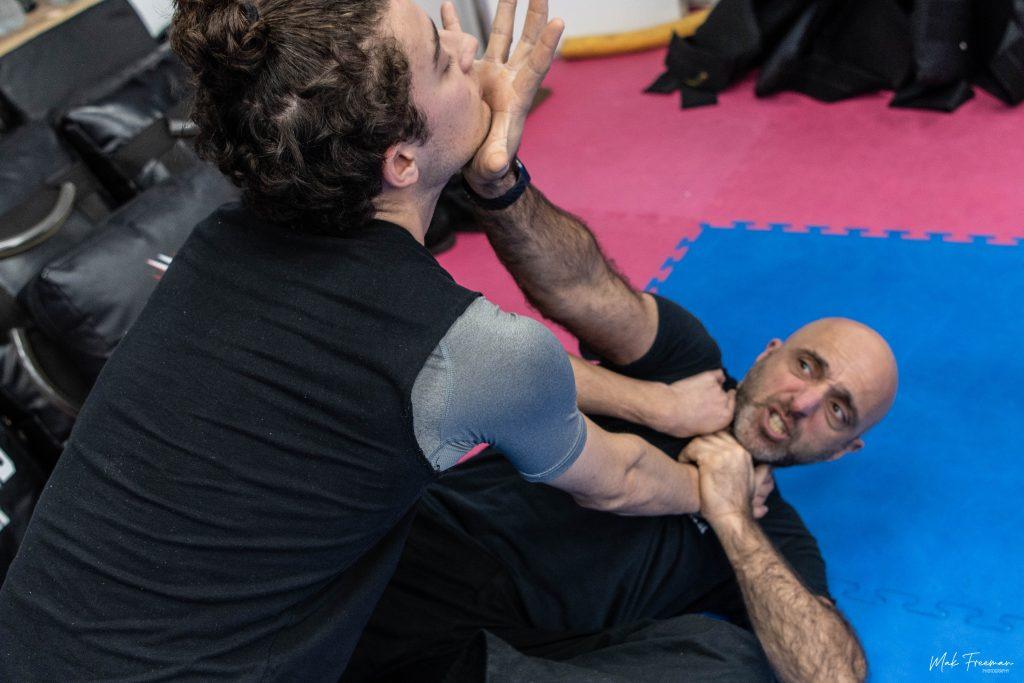 Image of two people doing krav maga, in a hold on the floor on a blue and pink mat.