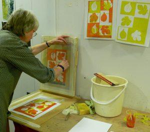 Picture of a woman using a screen printing press. Printing colourful images of flowers in red and green. She has a bucket, sponge and other printing materials next to her on the bench.